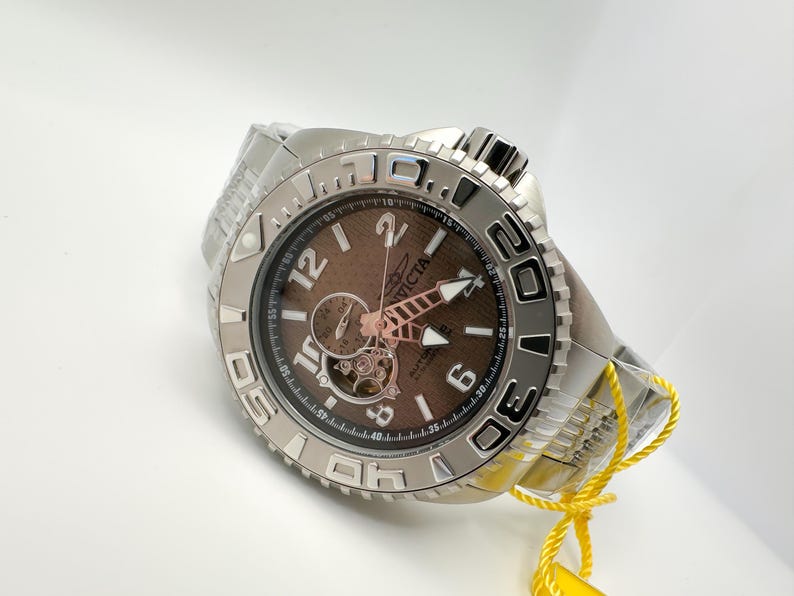 May include: Silver Invicta watch with a brown face and visible mechanical movement. The watch has a textured dial, luminous hands, and a stainless steel band. The watch has a rotating bezel with numerals and is new with a yellow tag.