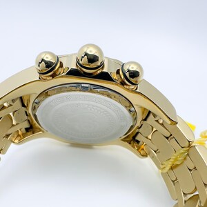 May include: A gold-toned wristwatch with a matching band. The watch features three prominent gold-colored buttons and a white face with text. The band is made of rectangular links. A yellow tag is attached to the band.