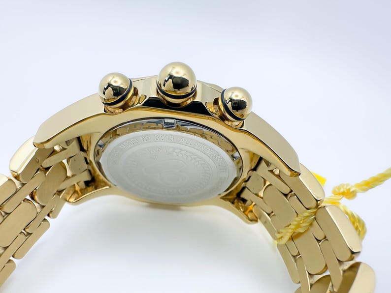 May include: A gold-toned wristwatch with a matching band. The watch features three prominent gold-colored buttons and a white face with text. The band is made of rectangular links. A yellow tag is attached to the band.