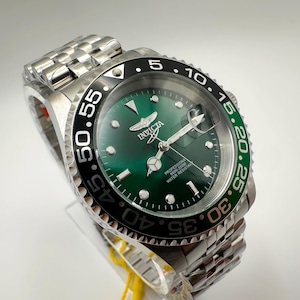 May include: A stainless steel Invicta watch with a green dial and a black and green bezel. The watch features a silver band and a detailed face with the Invicta logo. The watch is water resistant.