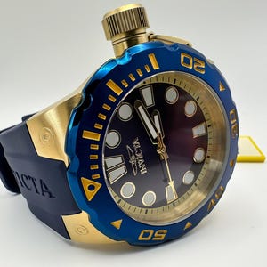 May include: A gold and blue Invicta watch with a dark blue face and a blue bezel. The watch has a gold crown and a dark blue rubber strap. The watch face has white and gold accents.