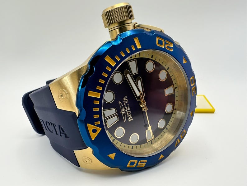 May include: A gold and blue Invicta watch with a dark blue face and a blue bezel. The watch has a gold crown and a dark blue rubber strap. The watch face has white and gold accents.