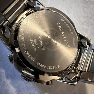 May include: Close-up of a stainless steel Caravelle watch. The watch back is visible, displaying the text "Designed by Bulova", "Water Resistant 100 meters / 330 feet", and other details. The watch band is also visible.
