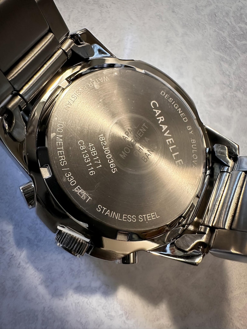 May include: Close-up of a stainless steel Caravelle watch. The watch back is visible, displaying the text "Designed by Bulova", "Water Resistant 100 meters / 330 feet", and other details. The watch band is also visible.