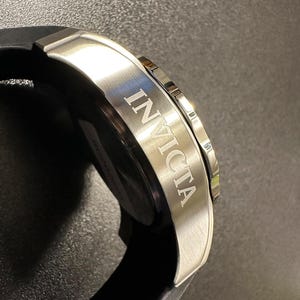 May include: A silver-toned stainless steel watch case with the word INVICTA engraved on the side. The watch features a black silicone strap and a metallic bezel with notched edges, showcasing a brushed finish on the side of the watch housing.