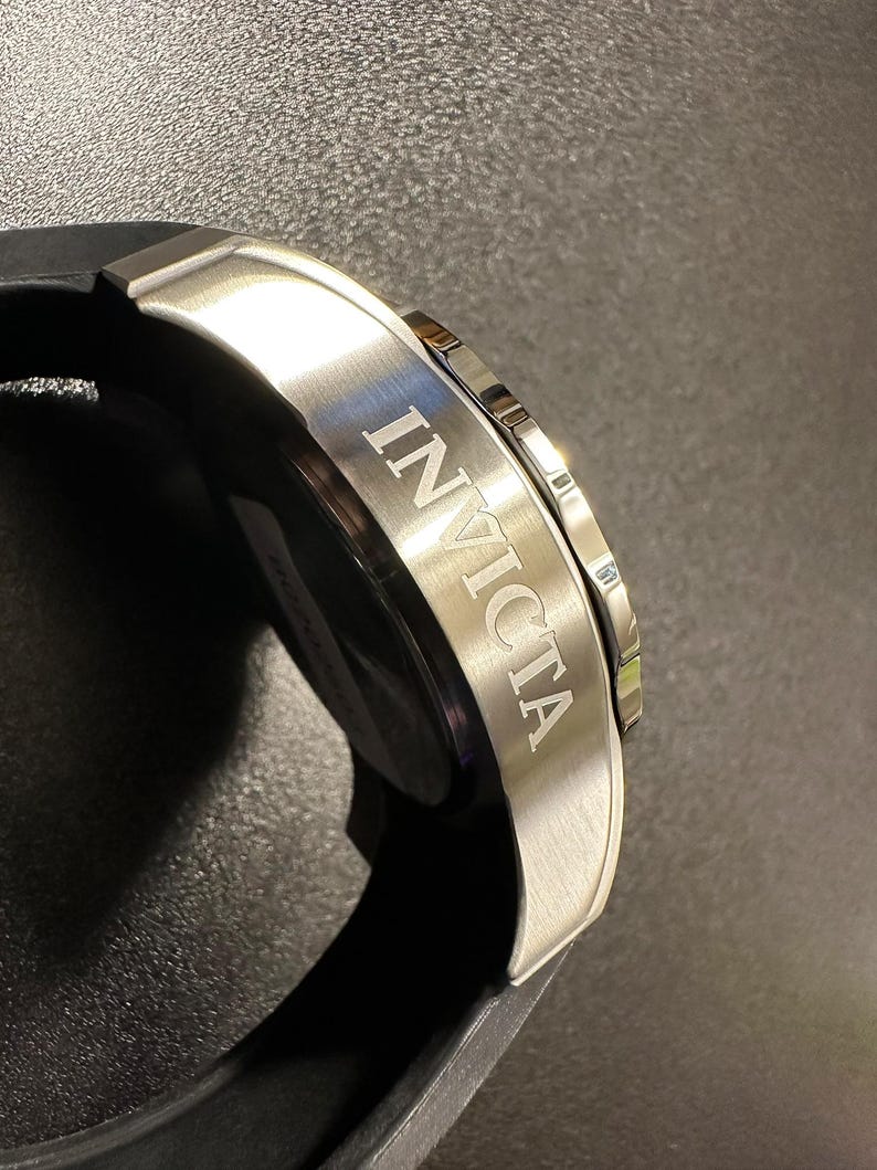 May include: A silver-toned stainless steel watch case with the word INVICTA engraved on the side. The watch features a black silicone strap and a metallic bezel with notched edges, showcasing a brushed finish on the side of the watch housing.