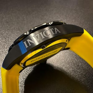 May include: An Invicta watch with a black case and a bright yellow strap. The word "INVICTA" is etched on the black bezel. The watch face is partially visible, with the number "6052509" printed on it.