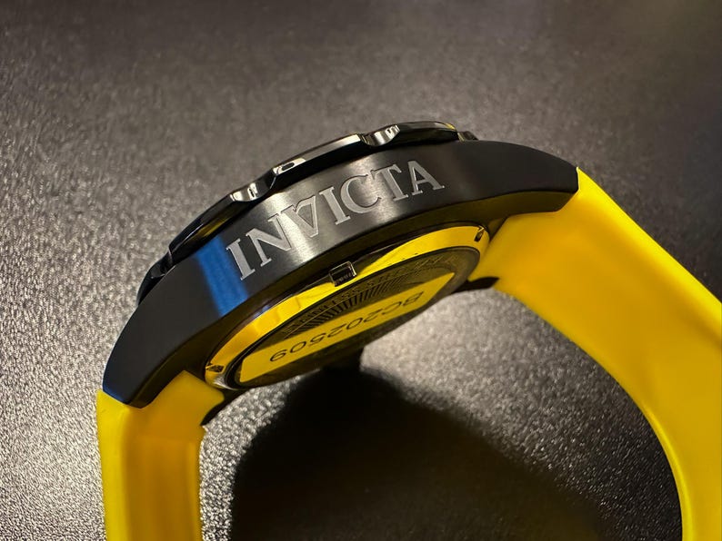 May include: An Invicta watch with a black case and a bright yellow strap. The word "INVICTA" is etched on the black bezel. The watch face is partially visible, with the number "6052509" printed on it.