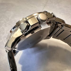 May include: A close-up of a silver-toned wristwatch with a stainless steel band. The watch face is partially visible, and the watch has two round buttons on the side. The band is made of rectangular links.