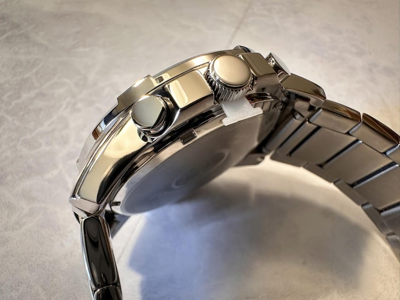 May include: A close-up of a silver-toned wristwatch with a stainless steel band. The watch face is partially visible, and the watch has two round buttons on the side. The band is made of rectangular links.