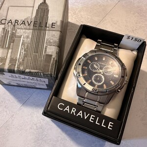May include: A Caravelle men's watch with a silver-tone stainless steel band and a black dial. The watch is displayed in a black box with the Caravelle logo and a cityscape design. The watch face has three sub-dials.