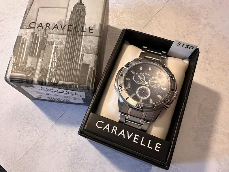 May include: A Caravelle men's watch with a silver-tone stainless steel band and a black dial. The watch is displayed in a black box with the Caravelle logo and a cityscape design. The watch face has three sub-dials.