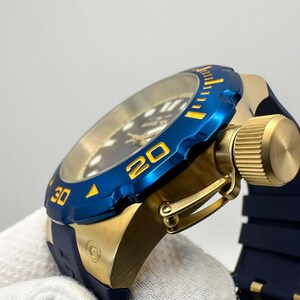 May include: A close-up of a gold-toned watch with a blue bezel and a dark blue band. The bezel has yellow markings at the 20 and 30-minute marks. The watch has a gold-toned crown and a dark blue rubber strap.