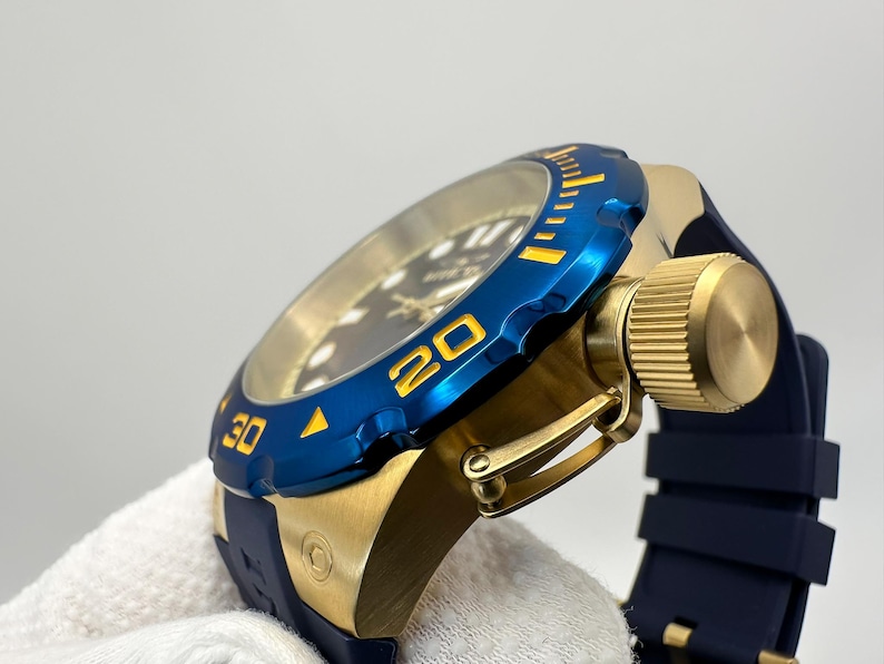 May include: A close-up of a gold-toned watch with a blue bezel and a dark blue band. The bezel has yellow markings at the 20 and 30-minute marks. The watch has a gold-toned crown and a dark blue rubber strap.