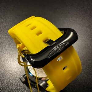 May include: A close-up of a yellow watch strap with a black clasp. The strap has rectangular cutouts and is secured with a black buckle. A yellow tag is attached to the watch.