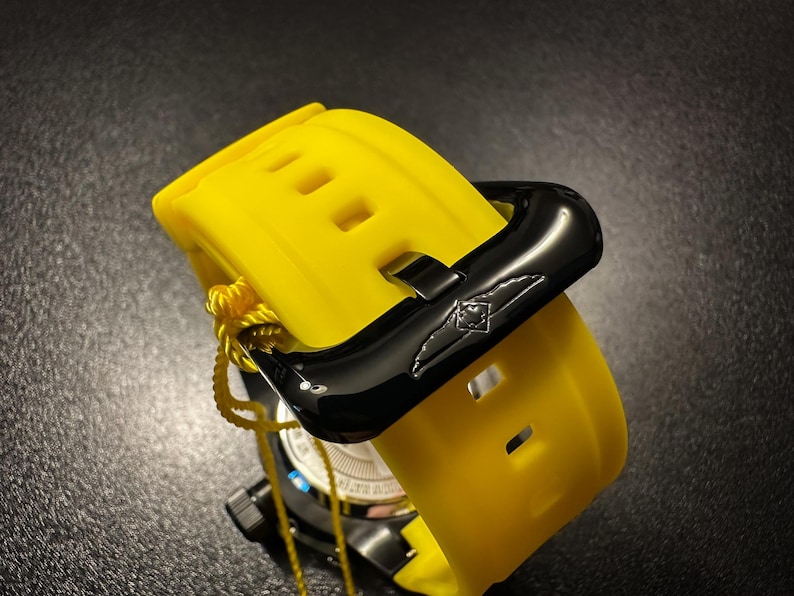 May include: A close-up of a yellow watch strap with a black clasp. The strap has rectangular cutouts and is secured with a black buckle. A yellow tag is attached to the watch.