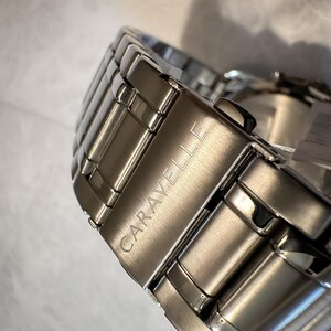 May include: Close-up of a silver-toned Caravelle watch band. The band is made of rectangular metal links, with the brand name engraved on the clasp. The watch band is highly reflective, with a polished finish.