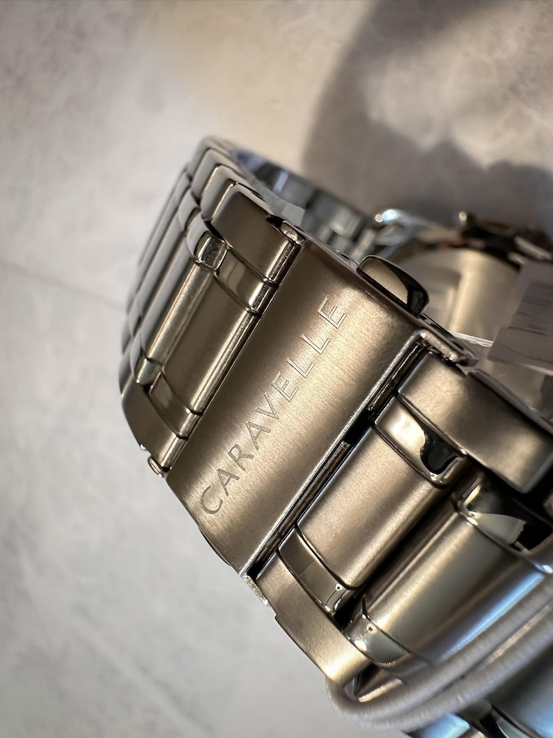 May include: Close-up of a silver-toned Caravelle watch band. The band is made of rectangular metal links, with the brand name engraved on the clasp. The watch band is highly reflective, with a polished finish.