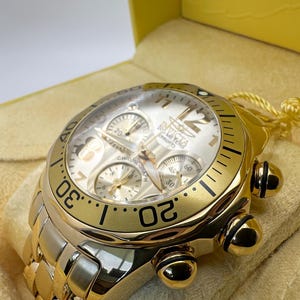 May include: A gold-toned Invicta chronograph watch with a white face, three sub-dials, and a gold bezel. The watch features a gold link bracelet and is presented in a yellow box with a cream-colored interior.