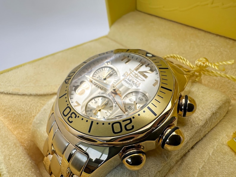 May include: A gold-toned Invicta chronograph watch with a white face, three sub-dials, and a gold bezel. The watch features a gold link bracelet and is presented in a yellow box with a cream-colored interior.