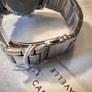 May include: A stainless steel watch with a silver band and clasp. The watch is new, with a tag attached. The brand name, Caravelle, is printed on the tag. The watch band is made of rectangular links.