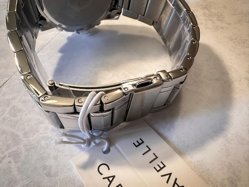 May include: A stainless steel watch with a silver band and clasp. The watch is new, with a tag attached. The brand name, Caravelle, is printed on the tag. The watch band is made of rectangular links.
