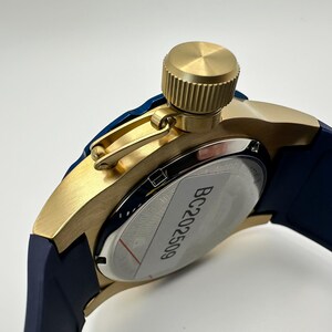 May include: Close-up of a gold-toned watch with a navy blue band. The watch features a textured crown and a silver-colored back with the text "BC202509". The watch has a modern, sporty design.