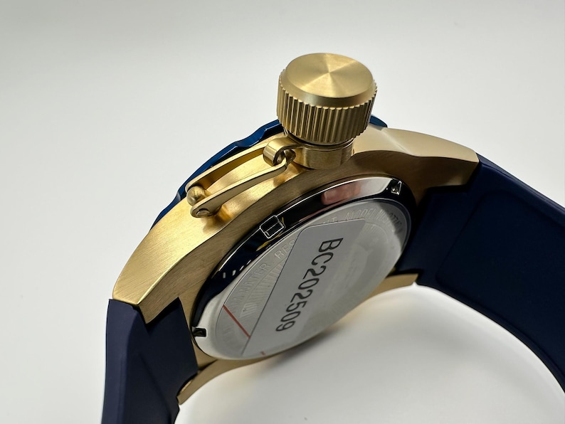 May include: Close-up of a gold-toned watch with a navy blue band. The watch features a textured crown and a silver-colored back with the text "BC202509". The watch has a modern, sporty design.