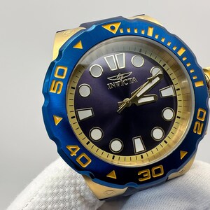 May include: A blue and gold Invicta watch with a dark blue face. The watch has gold hands and markers, and a blue rotating bezel with gold numerals. The watch is held by a gloved hand.