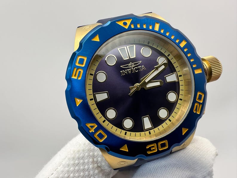 May include: A blue and gold Invicta watch with a dark blue face. The watch has gold hands and markers, and a blue rotating bezel with gold numerals. The watch is held by a gloved hand.