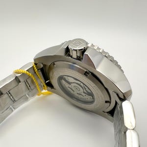 May include: A silver stainless steel wristwatch with a silver strap and a yellow tag. The watch has a round face with a visible mechanism and a textured bezel. The watch is angled, showing the side and back.