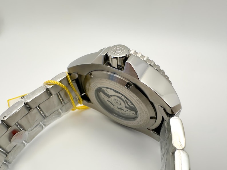 May include: A silver stainless steel wristwatch with a silver strap and a yellow tag. The watch has a round face with a visible mechanism and a textured bezel. The watch is angled, showing the side and back.