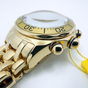 May include: Close-up of a gold-toned wristwatch with a clear, domed crystal. The watch features a black bezel with white numerals and a matching gold link band. A yellow tag hangs from the watch.