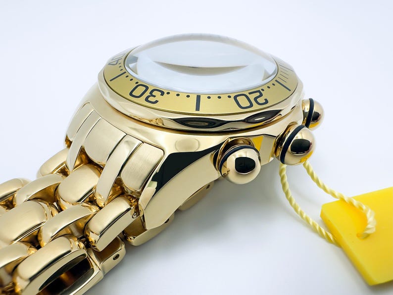 May include: Close-up of a gold-toned wristwatch with a clear, domed crystal. The watch features a black bezel with white numerals and a matching gold link band. A yellow tag hangs from the watch.