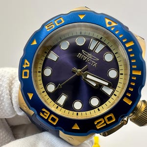 May include: A close-up of an Invicta watch with a blue and gold bezel. The watch face is a deep blue with gold hands and markers. The watch has a large crown and is held by a gloved hand.