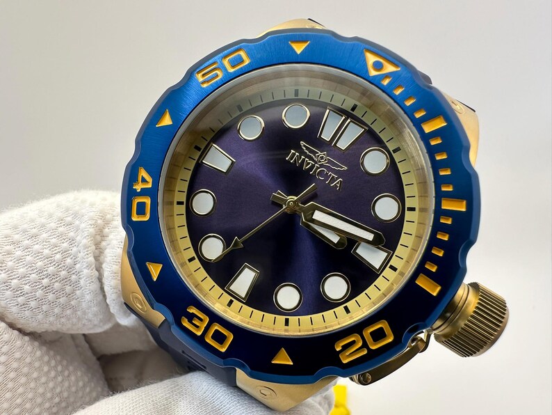 May include: A close-up of an Invicta watch with a blue and gold bezel. The watch face is a deep blue with gold hands and markers. The watch has a large crown and is held by a gloved hand.