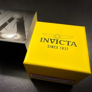 May include: A bright yellow box with the brand name "INVICTA" and the text "SINCE 1837" printed in black. A black and silver box is partially visible in the background. The boxes are on a dark surface.