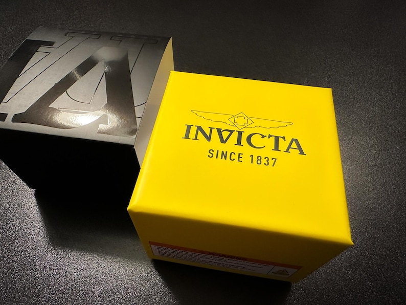May include: A bright yellow box with the brand name "INVICTA" and the text "SINCE 1837" printed in black. A black and silver box is partially visible in the background. The boxes are on a dark surface.