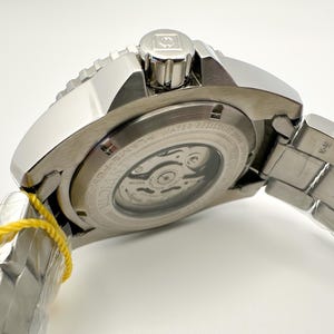 May include: A close-up of a silver Invicta watch. The watch has a visible movement through the back, a textured bezel, and a metal strap. The watch is new, with a yellow tag attached.