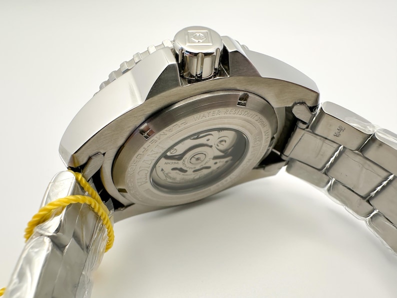 May include: A close-up of a silver Invicta watch. The watch has a visible movement through the back, a textured bezel, and a metal strap. The watch is new, with a yellow tag attached.