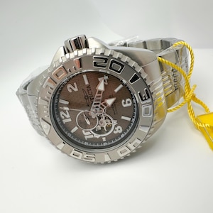 May include: A silver Invicta watch with a brown face and a stainless steel strap. The watch has a rotating bezel with white numerals and a yellow tag attached.