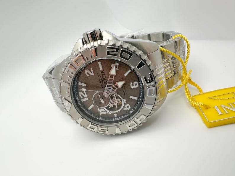 May include: A silver Invicta watch with a brown face and a stainless steel strap. The watch has a rotating bezel with white numerals and a yellow tag attached.