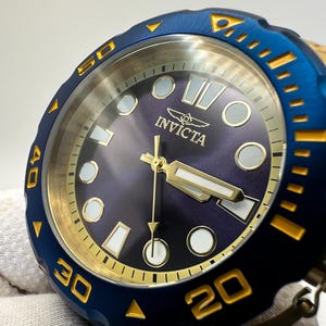 May include: Close-up of an Invicta watch with a blue bezel and gold accents. The watch face is a deep purple with gold hands and markers. The watch has a textured blue bezel with gold numerals and markers. The Invicta logo is visible on the watch face.
