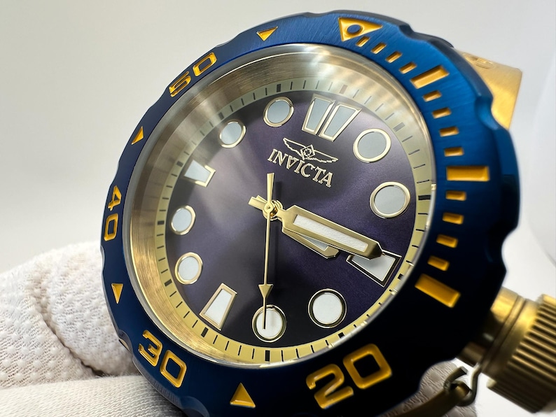 May include: Close-up of an Invicta watch with a blue bezel and gold accents. The watch face is a deep purple with gold hands and markers. The watch has a textured blue bezel with gold numerals and markers. The Invicta logo is visible on the watch face.