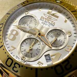 May include: Gold-toned Invicta chronograph watch with a silver face. The watch features three sub-dials, gold hands, and large gold numerals. The watch is water-resistant to 200 meters (650 feet). The watch has the word "CHRONOGRAPH" on the face.