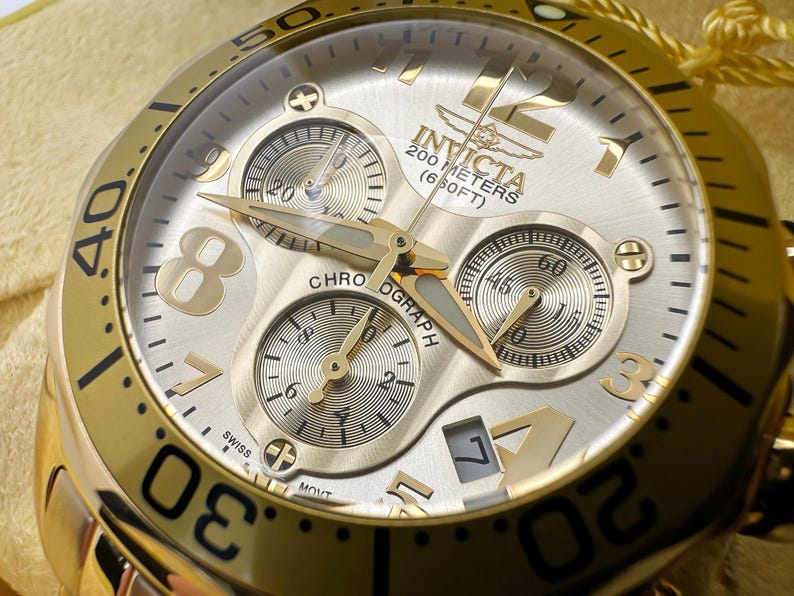 May include: Gold-toned Invicta chronograph watch with a silver face. The watch features three sub-dials, gold hands, and large gold numerals. The watch is water-resistant to 200 meters (650 feet). The watch has the word "CHRONOGRAPH" on the face.