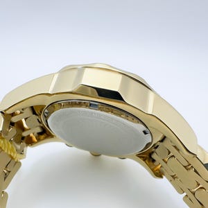 May include: Close-up of a gold-toned wristwatch. The watch features a gold band and a gold case. The watch face is white with black text. The watch is angled, showing the underside and the band.