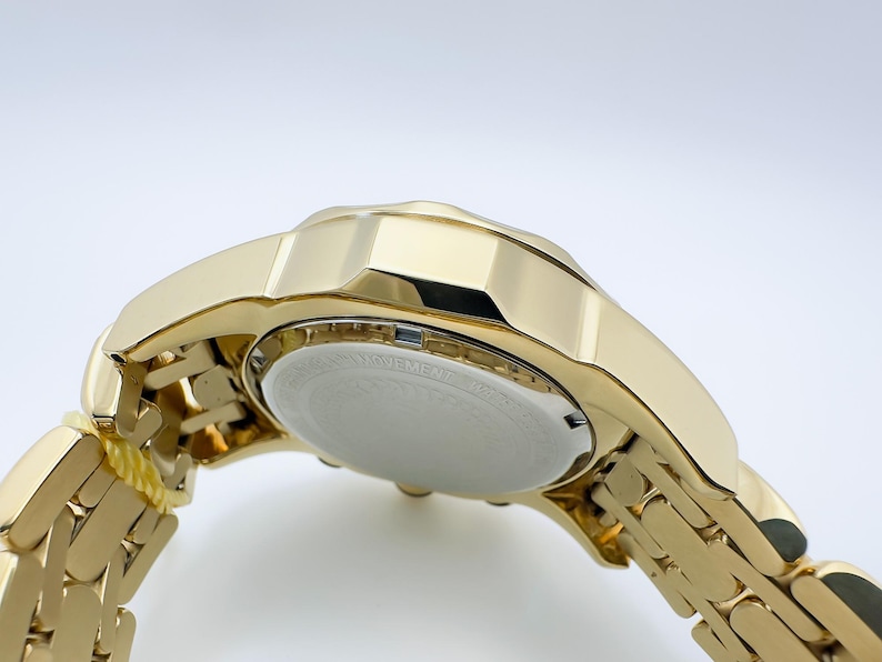 May include: Close-up of a gold-toned wristwatch. The watch features a gold band and a gold case. The watch face is white with black text. The watch is angled, showing the underside and the band.