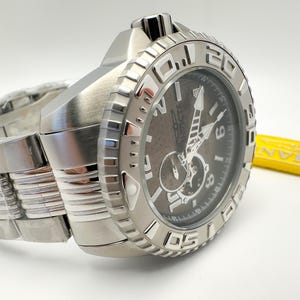 May include: A stainless steel Invicta watch with a dark grey face and silver hands. The watch has a large, textured bezel with white numerals and a matching stainless steel band. The watch is new and still has the protective plastic on the band.