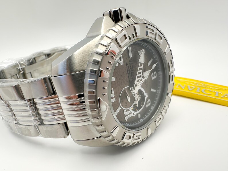 May include: A stainless steel Invicta watch with a dark grey face and silver hands. The watch has a large, textured bezel with white numerals and a matching stainless steel band. The watch is new and still has the protective plastic on the band.